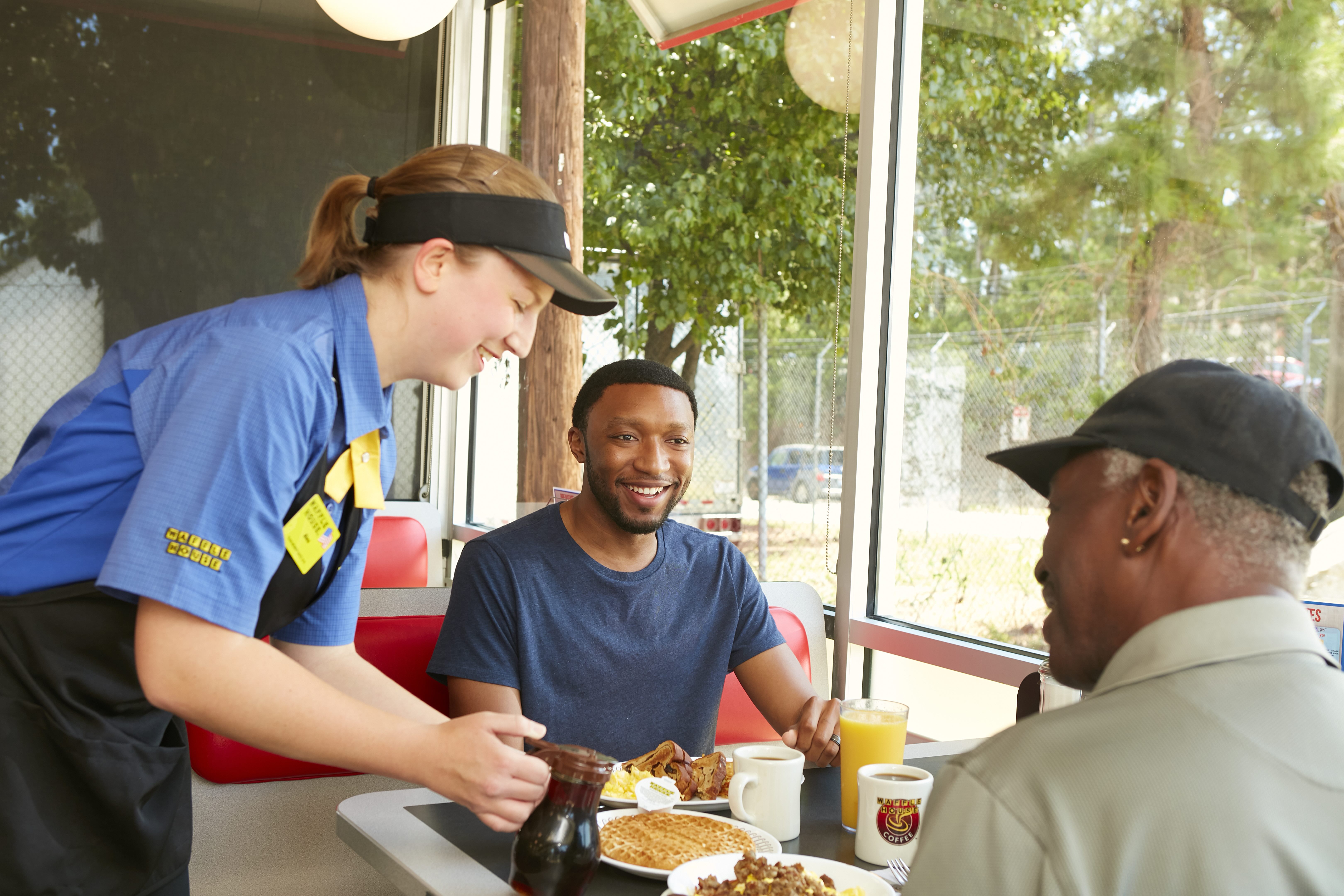 Smiling Waffle House employee serving a waffle and syrup to two smiling customers